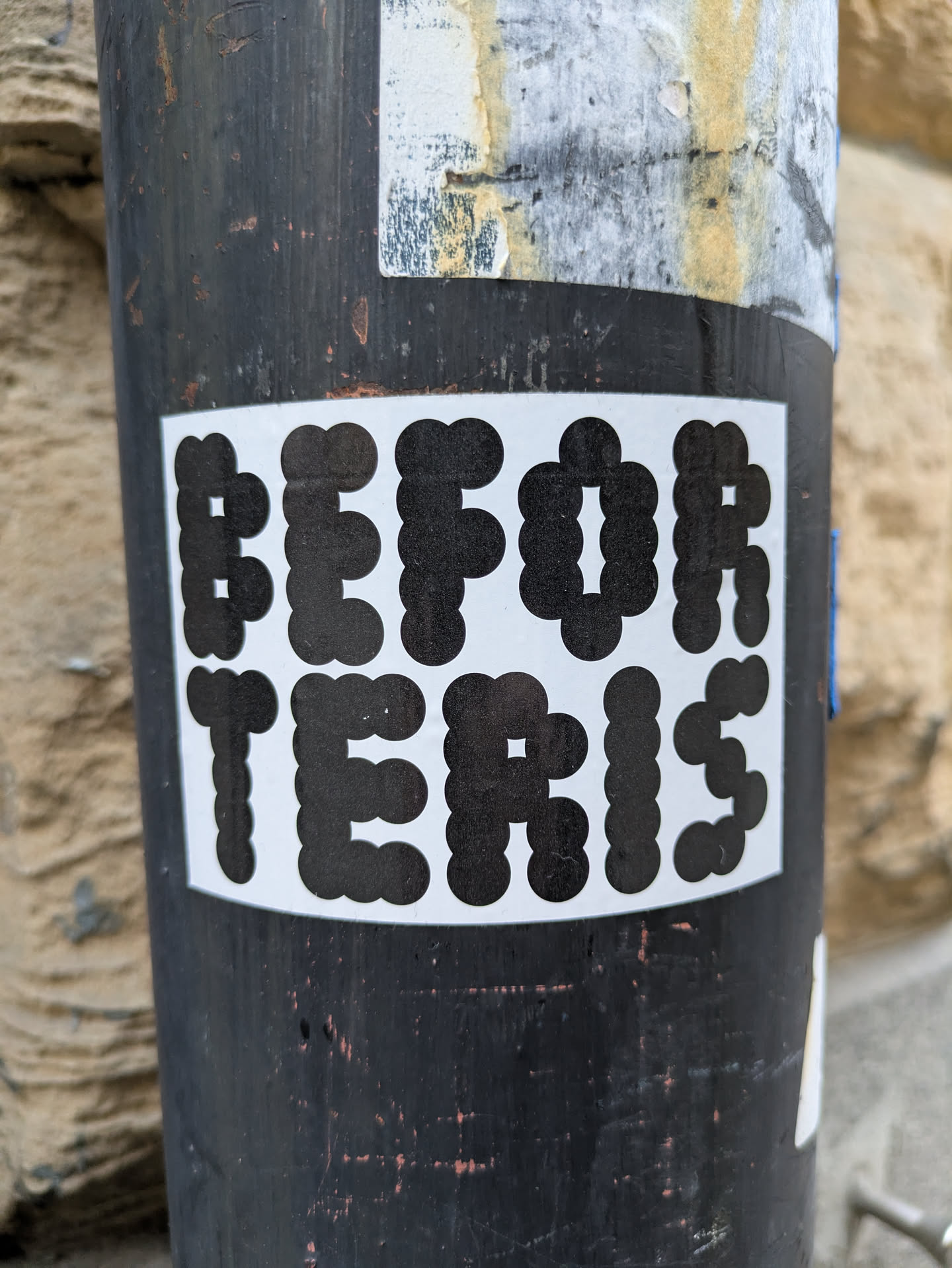A black and white sticker with the text "BEFORE TETRIS" in a pixelated font. The sticker is attached to a dark-colored metal pole.