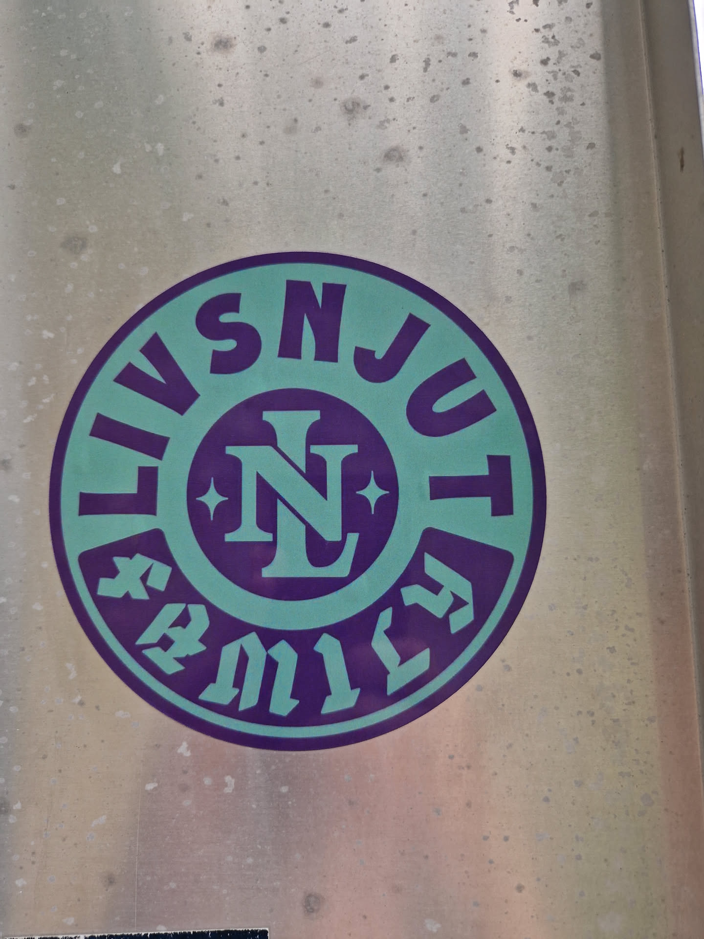 Close-up of a round sticker with the text "LIVSNJUT" circling a central logo featuring the letters "NL" within a stylized emblem. The sticker has a teal and purple color scheme and appears to be affixed to a metallic surface.