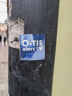 A blue square sticker affixed to a dark-colored pole. The sticker features the text 'Q-TIE alert!' in white, with a small pink heart symbol following the exclamation mark.  The bottom of the sticker includes the logo 'Q SUMMIT'.