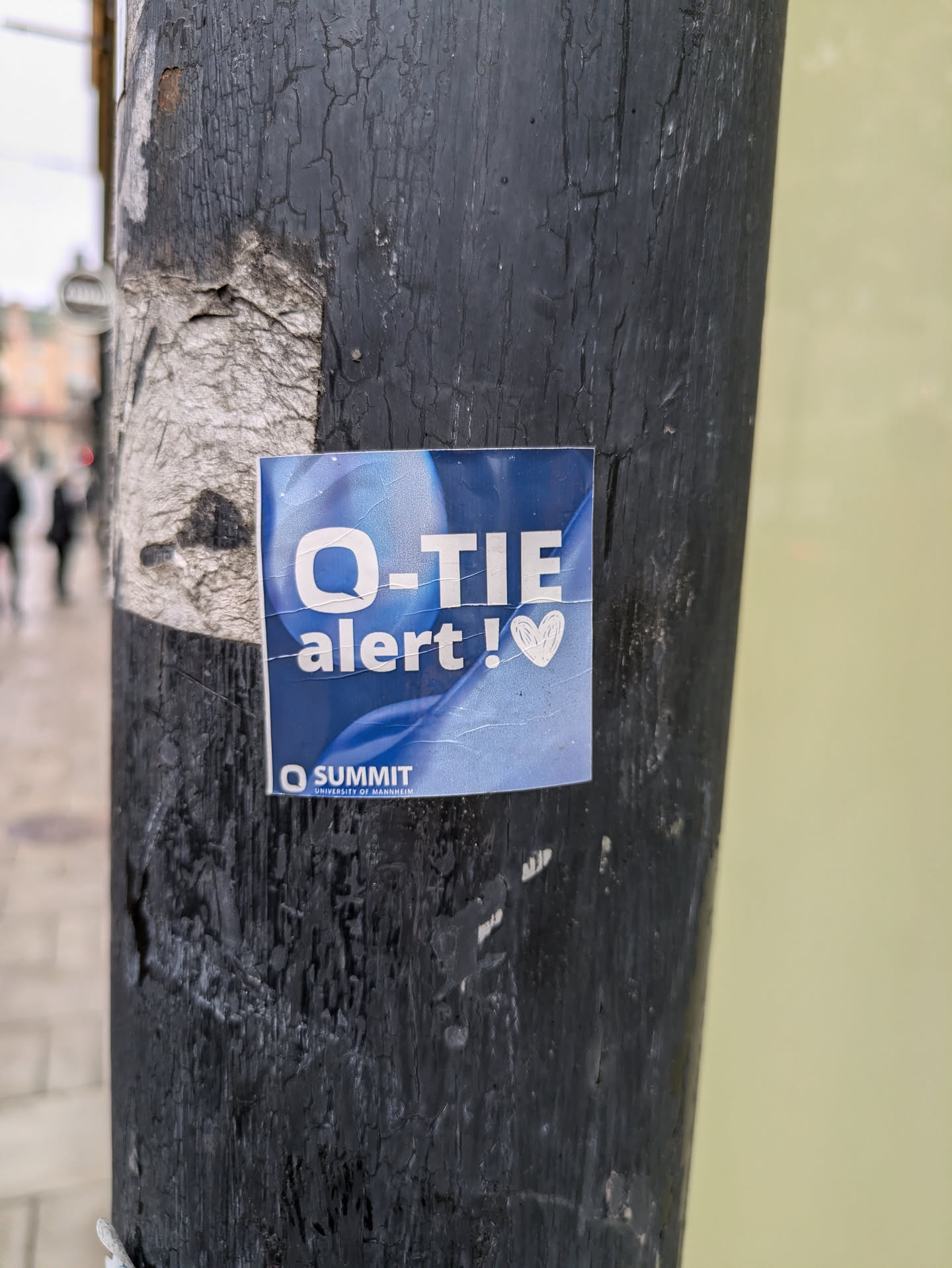 A blue square sticker affixed to a dark-colored pole. The sticker features the text 'Q-TIE alert!' in white, with a small pink heart symbol following the exclamation mark.  The bottom of the sticker includes the logo 'Q SUMMIT'.