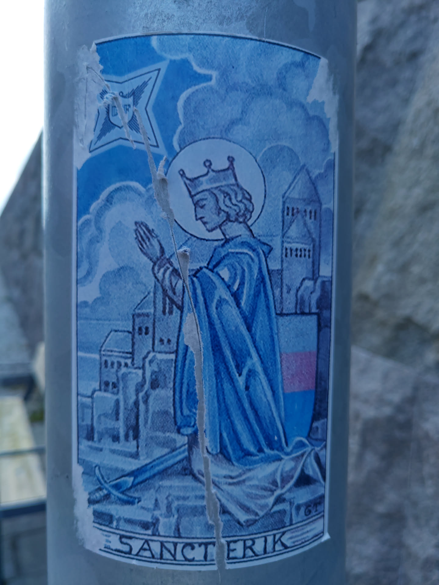 A sticker depicting a medieval illustration of Sanct Erik, a crowned figure kneeling in prayer. The image is in shades of blue and features a city landscape in the background.  The sticker shows some damage from wear and tear.