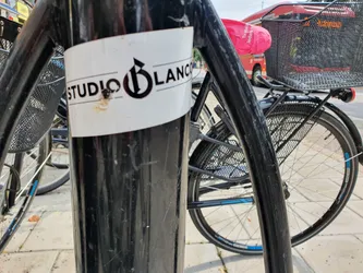 A close-up shot of a Studio Blanc sticker affixed to a black pole. The sticker is white with black text and logo. The logo appears to be a stylized 'B'.  Some minor damage is visible on the sticker.