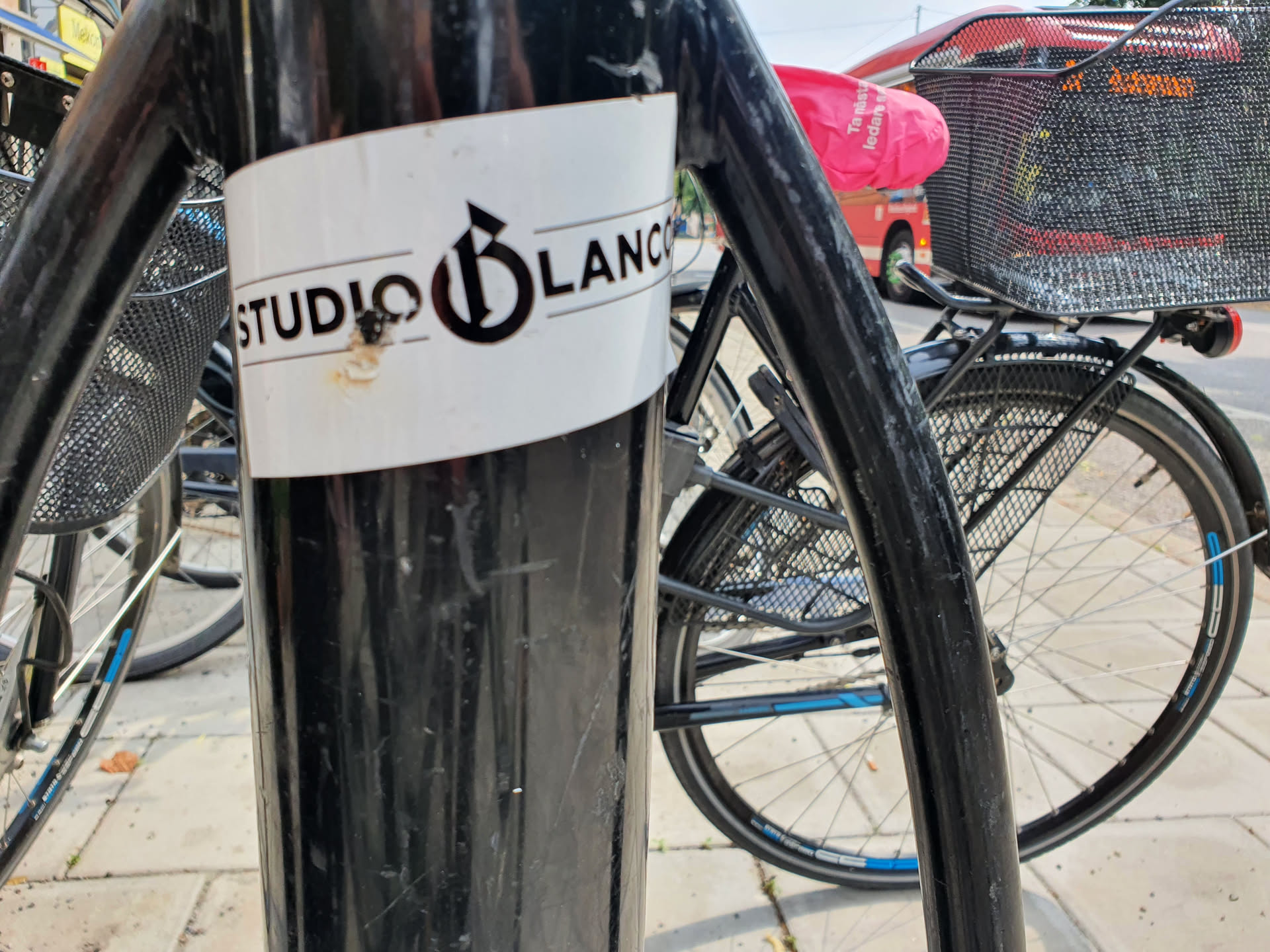 A close-up shot of a Studio Blanc sticker affixed to a black pole. The sticker is white with black text and logo. The logo appears to be a stylized 'B'.  Some minor damage is visible on the sticker.