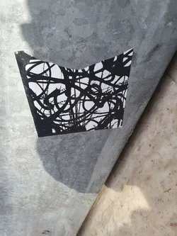 A sticker with an abstract design featuring black ink lines on a white background. The lines are chaotic and interwoven, creating a sense of movement and energy. The sticker is attached to a metallic surface.