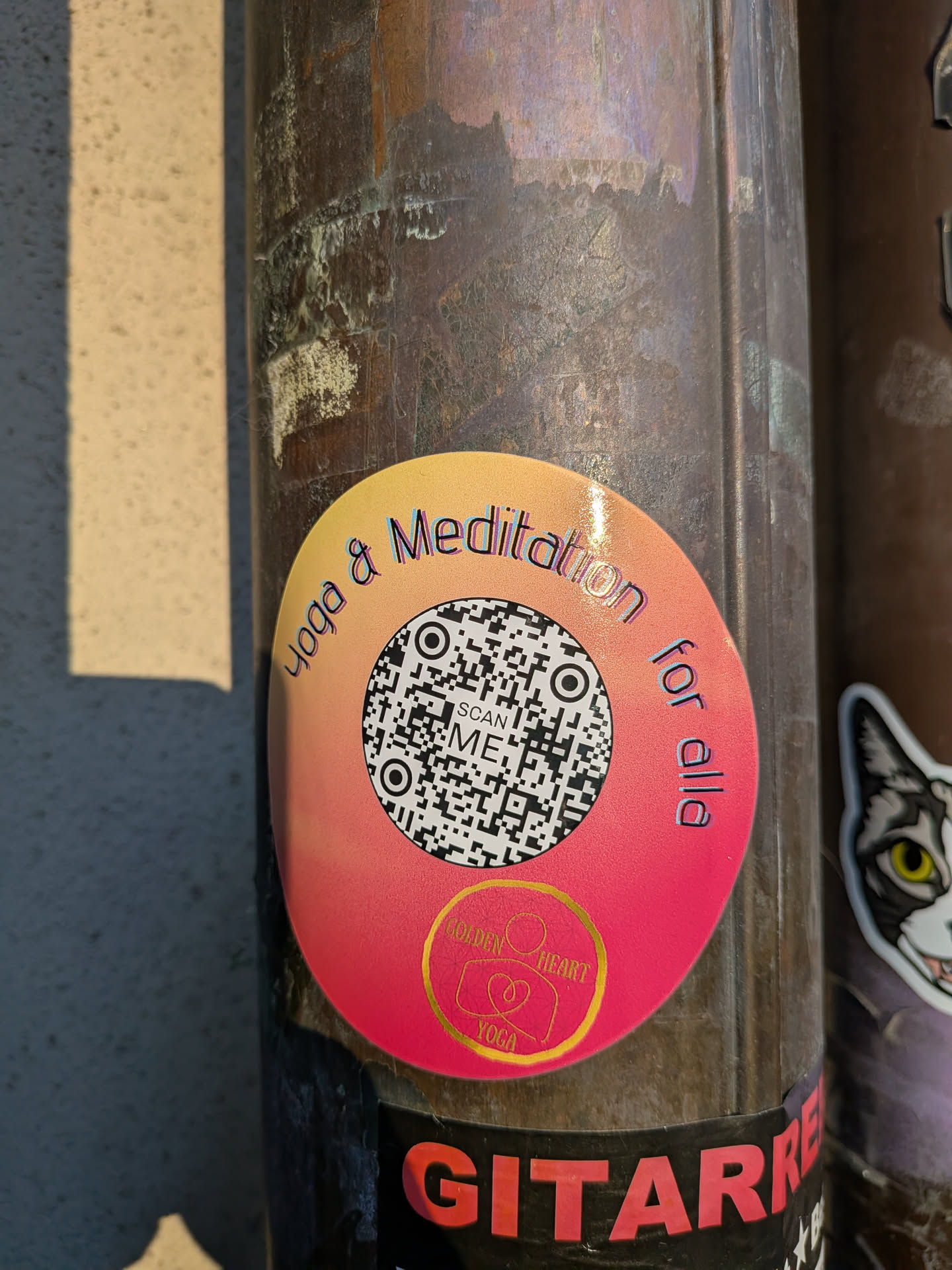 A circular sticker featuring a vibrant gradient background, transitioning from a light orange-yellow at the top to a rich magenta-pink at the bottom. A prominent black and white QR code occupies the center, with the text 'SCAN ME' clearly visible within it. Arching along the top edge of the sticker is the text 'Yoga & Meditation' in a dark, slightly shadowed font. Along the bottom edge, curving upwards, is 'for alla'. At the very bottom center, within the pink area, is a small golden circular logo containing the text 'GOLDEN HEART YOGA' and a stylized icon depicting a person in a meditative pose with a heart shape.