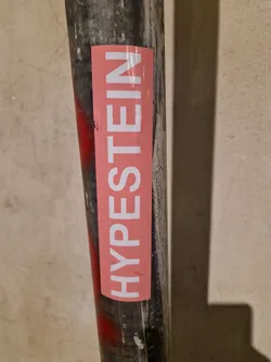 A pink sticker with the word "HYPESTEIN" written in white uppercase letters. The sticker is attached to a dark-grey metal pipe.