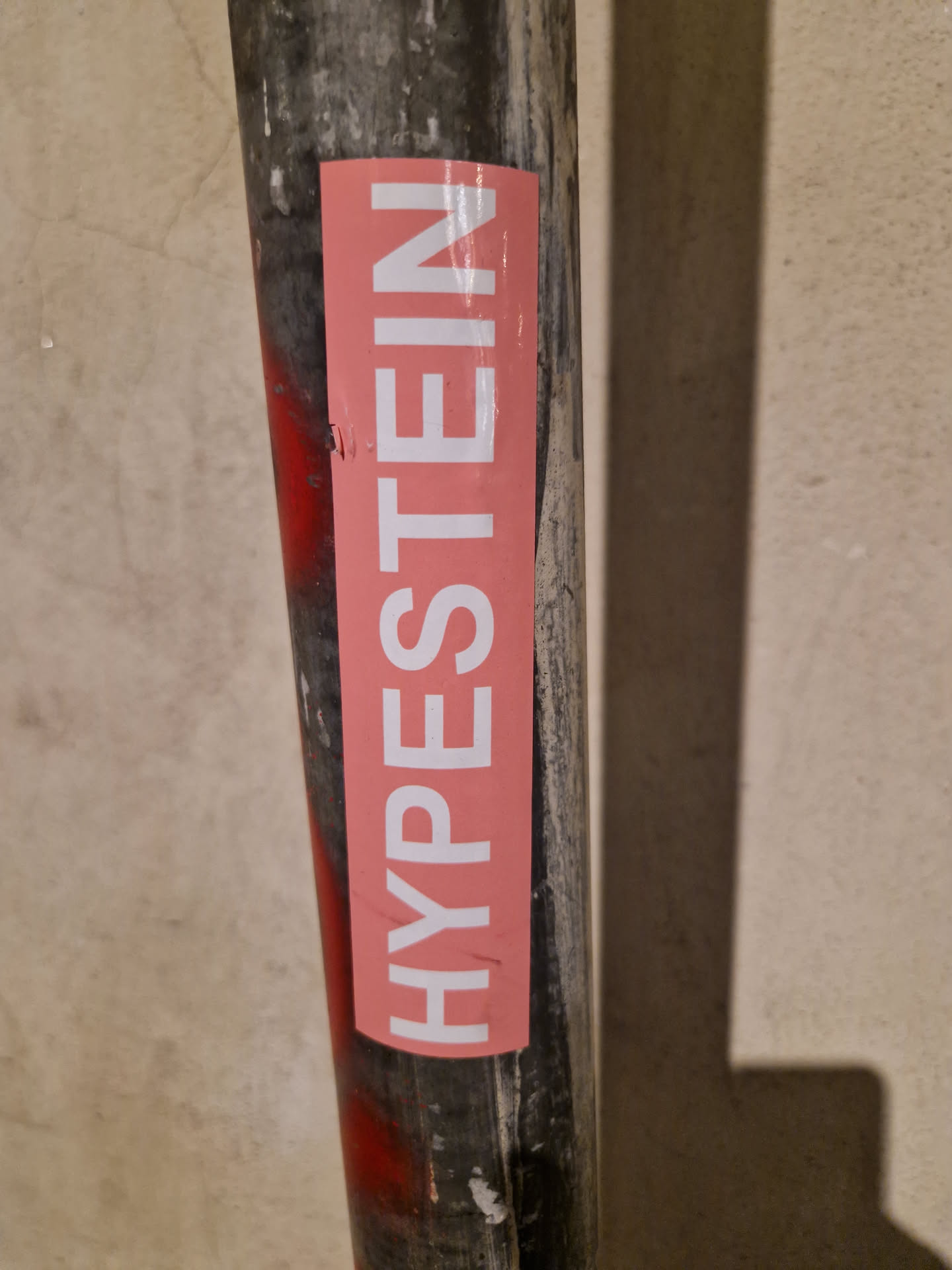 A pink sticker with the word "HYPESTEIN" written in white uppercase letters. The sticker is attached to a dark-grey metal pipe.