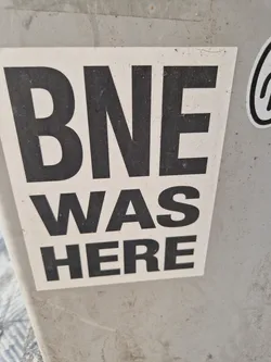 A simple sticker with the text "BNE WAS HERE" in bold black letters on a white background. The sticker appears to be affixed to a gray surface.