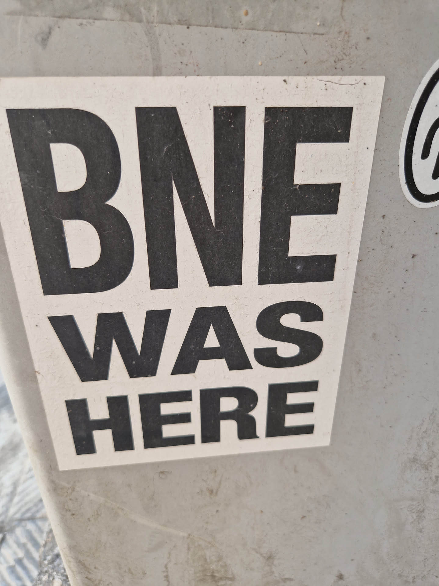A simple sticker with the text "BNE WAS HERE" in bold black letters on a white background. The sticker appears to be affixed to a gray surface.
