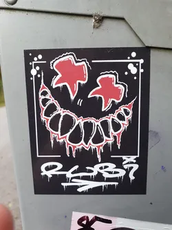A black and white sticker with a red creepy smile and two red stars on top.  The sticker has some graffiti-style text at the bottom that looks like a signature or tag. The style is reminiscent of edgy, alternative art.