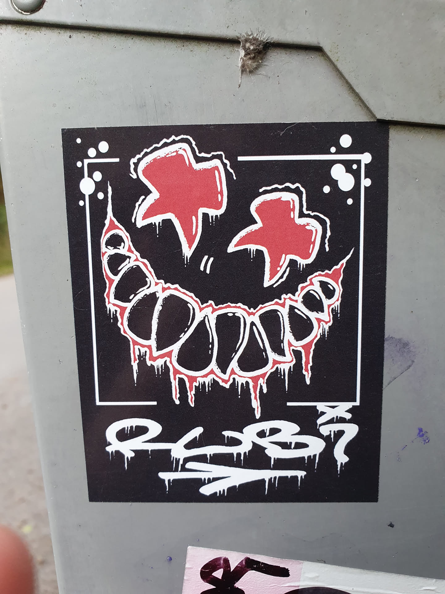 A black and white sticker with a red creepy smile and two red stars on top.  The sticker has some graffiti-style text at the bottom that looks like a signature or tag. The style is reminiscent of edgy, alternative art.