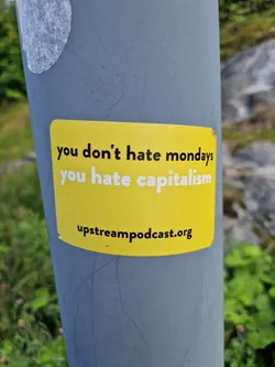 A yellow sticker with a political message on it. It is pasted on a gray pole outdoors. The text on the sticker says "you don't hate mondays you hate capitalism" and also includes a website address.