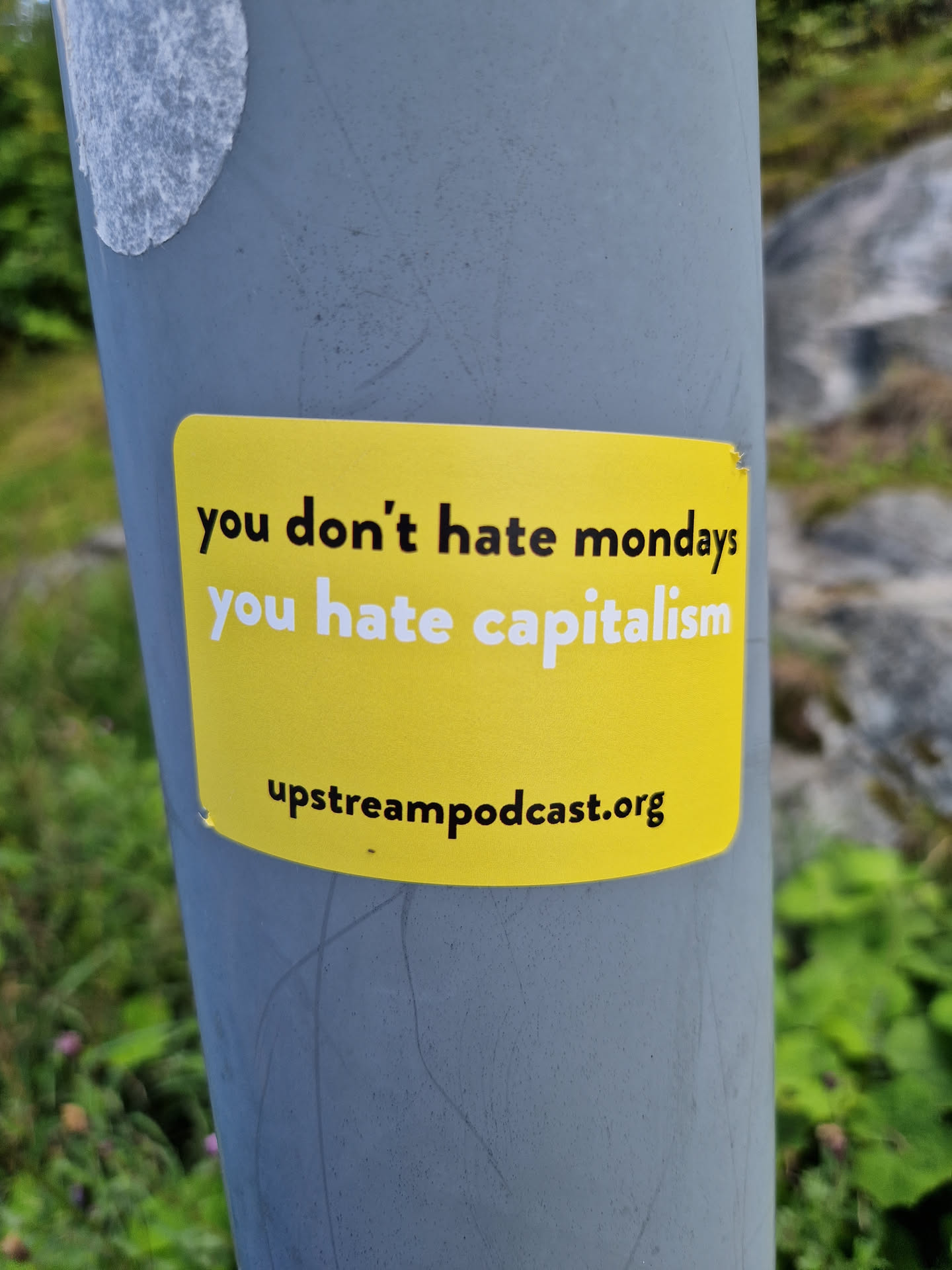 A yellow sticker with a political message on it. It is pasted on a gray pole outdoors. The text on the sticker says "you don't hate mondays you hate capitalism" and also includes a website address.