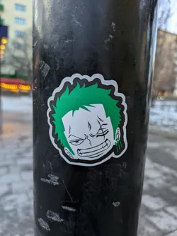 A sticker of Zoro from One Piece is stuck on a metal pole. He has green hair, is missing his left eye, and is wearing green earrings.