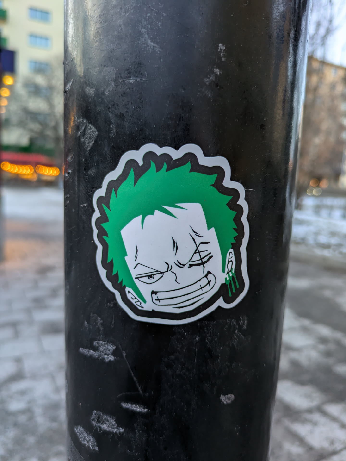 A sticker of Zoro from One Piece is stuck on a metal pole. He has green hair, is missing his left eye, and is wearing green earrings.