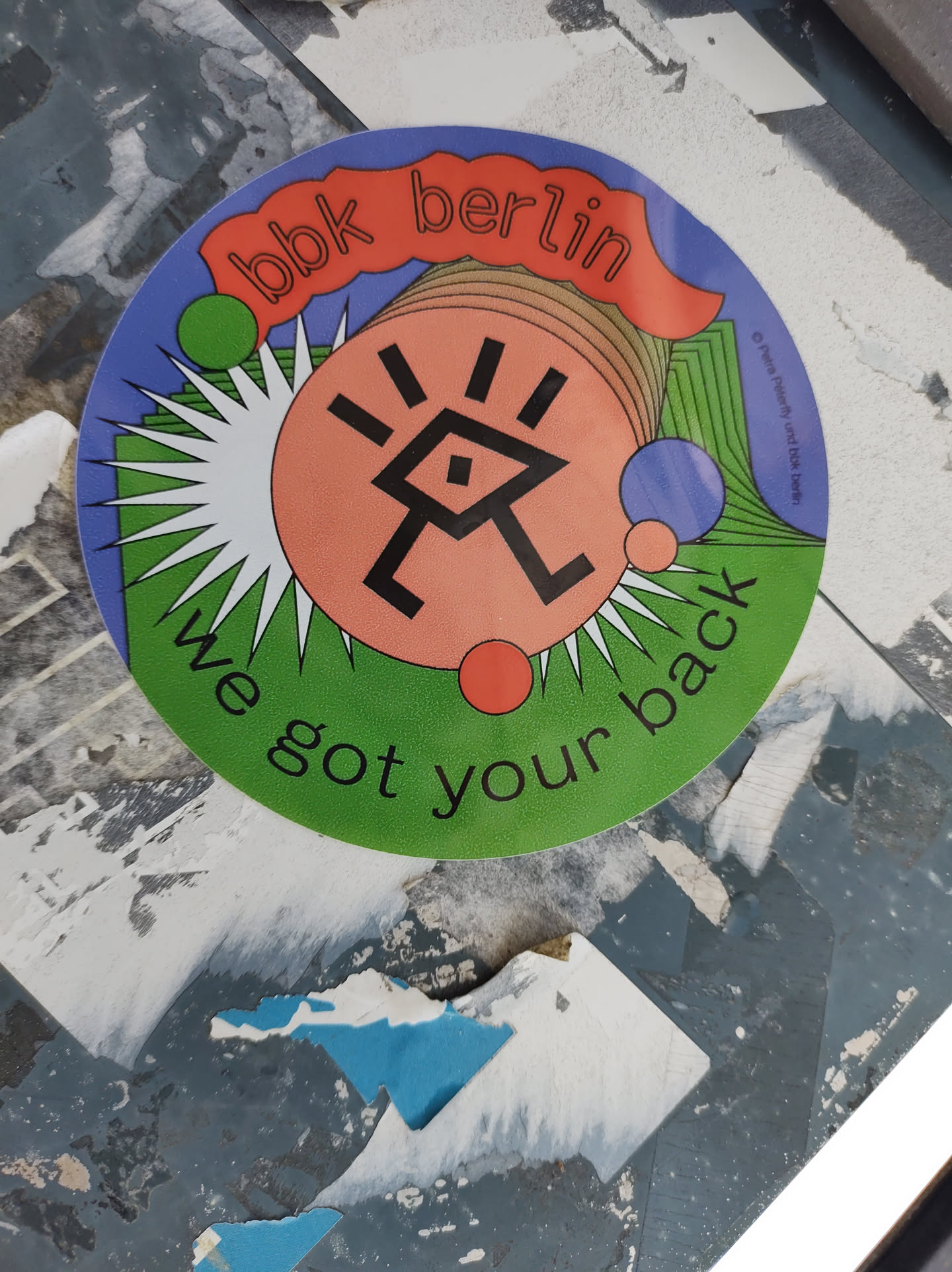 Round sticker with bbk berlin text and a logo. The logo is a stylized symbol with geometric shapes and lines. The colors are bright and bold, including purple, green, orange, and pink. The text "We got your back" is prominently displayed below the logo.