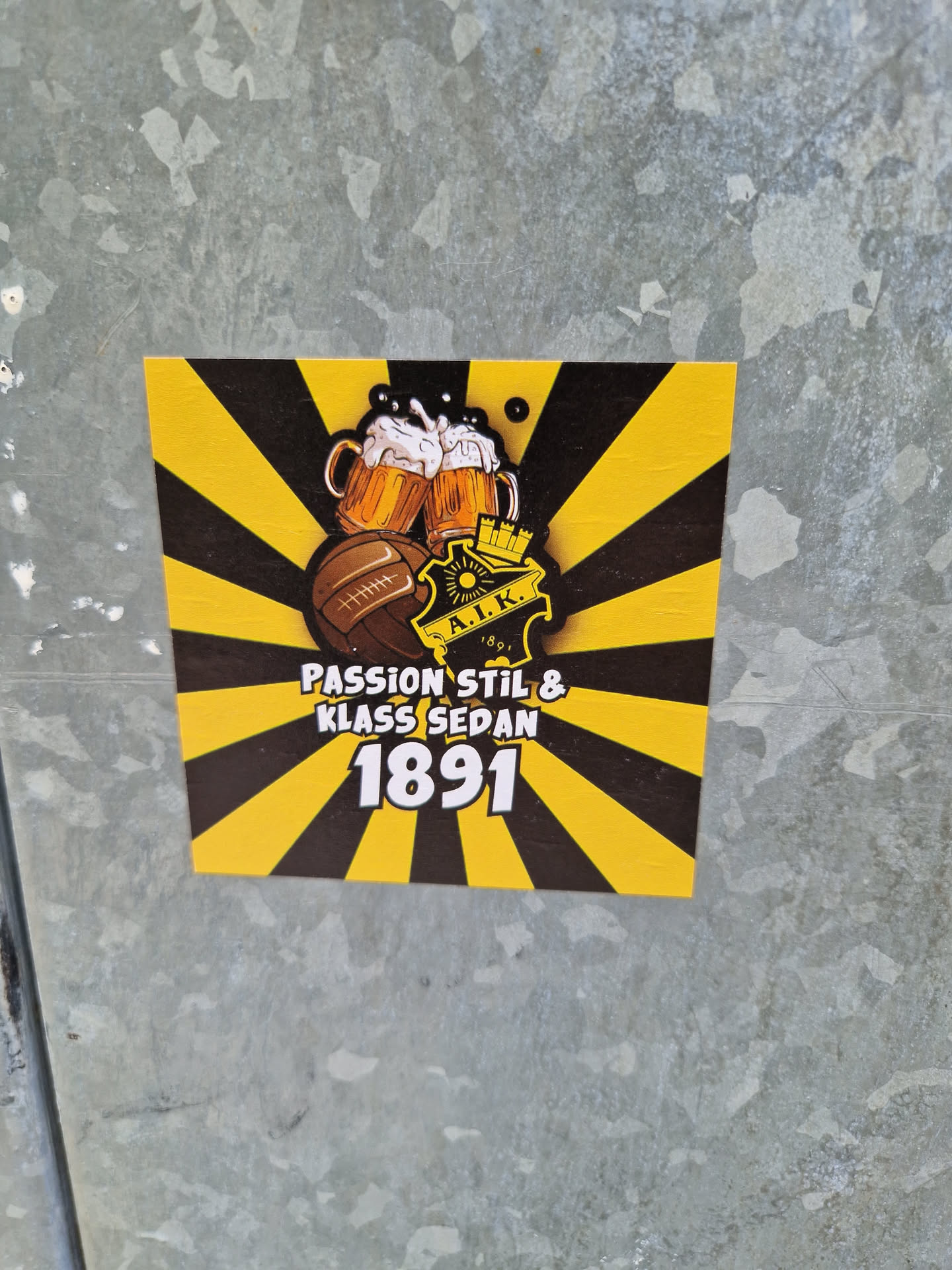 A yellow and black sticker with two beer steins, a soccer ball and the text "Passion Stil & Klass Sedan 1891".