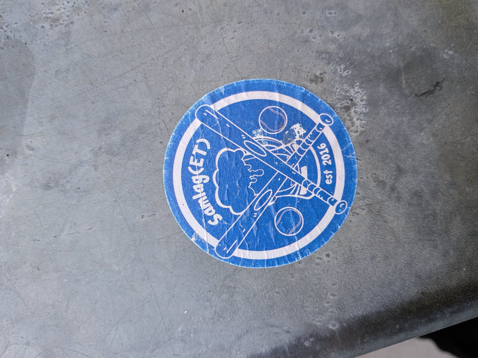 The sticker is circular with a blue background and a white border. In the center, two crossed baseball bats are depicted, with a baseball above them. A stylized cloud-like shape is visible behind the bats. Text is present along the circumference: 'Samlag(ET)' on the left and 'est 2016' on the right. The overall design is simple and graphic, suggesting a sports team or organization.