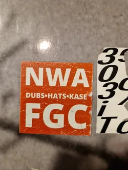 An orange sticker with the text NWA, DUBS.HATS.KASE, and FGC. The sticker appears worn and slightly damaged.
