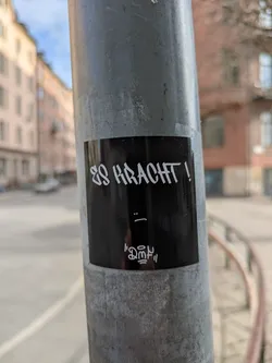 The image shows a black rectangular sticker adhered to a metal pole. The text "ES KRACHT!" is written in a stylized, somewhat angular white font. Underneath the text is a simple "_:" emoticon and a stylized artistic element. The sticker is placed on a pole in an urban setting.