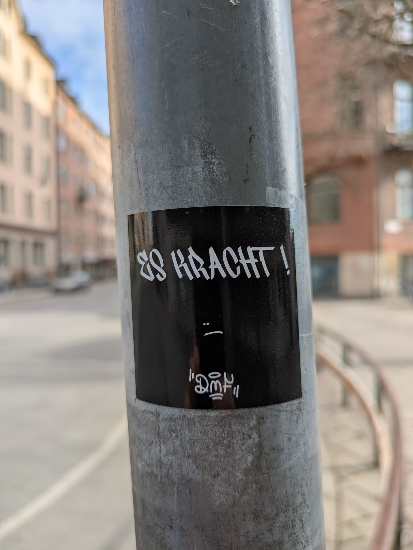 The image shows a black rectangular sticker adhered to a metal pole. The text "ES KRACHT!" is written in a stylized, somewhat angular white font. Underneath the text is a simple "_:" emoticon and a stylized artistic element. The sticker is placed on a pole in an urban setting.