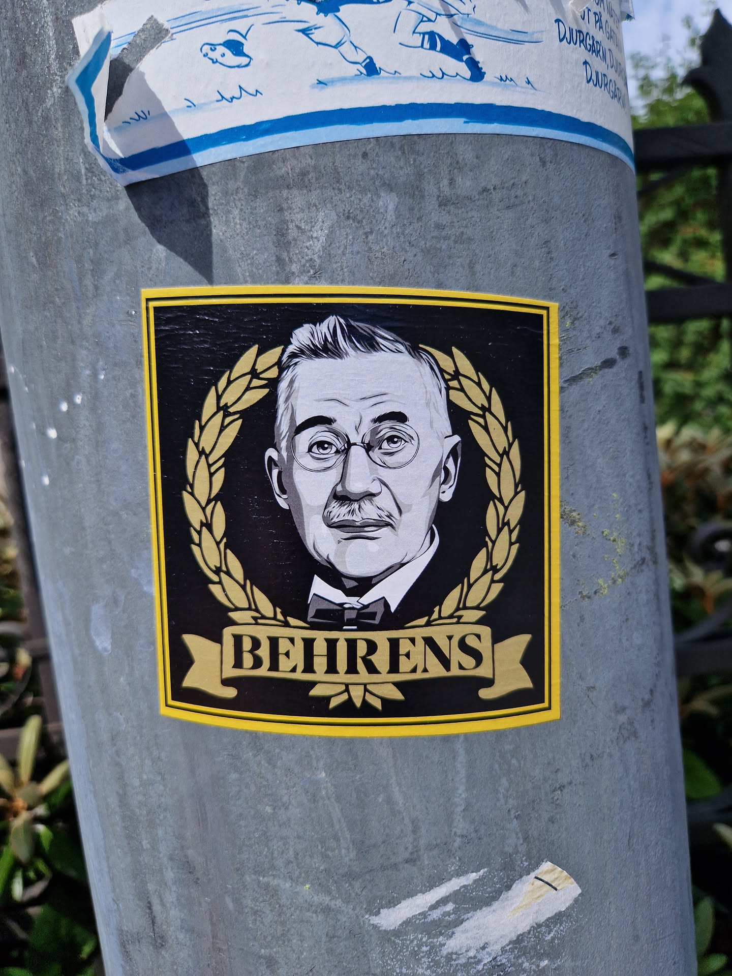 A black and gold sticker depicting Peter Behrens, a German architect and designer. The sticker features a portrait of Behrens surrounded by a laurel wreath, with his name written in a banner below. 