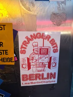 A white square sticker with red ink. It features a chef using a modified Gameboy to play chiptune music, positioned between two speakers. Text includes "STRÄÄNGER BITS" around the top, "BERLIN" at the bottom, and a Telegram link. The QR code on the gameboy screen leads to a URL related to chiptune music or the artist.