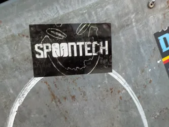 The sticker is rectangular with a black background. The text 'SPOONTECH' is prominently displayed in a bold, white, sans-serif font. The letters have a slightly distressed or worn appearance. There are thin, sketchy white lines and circular shapes around the text, suggesting a dynamic or technical design. The sticker appears to be adhered to a metallic surface with a visible grain and some rust spots.