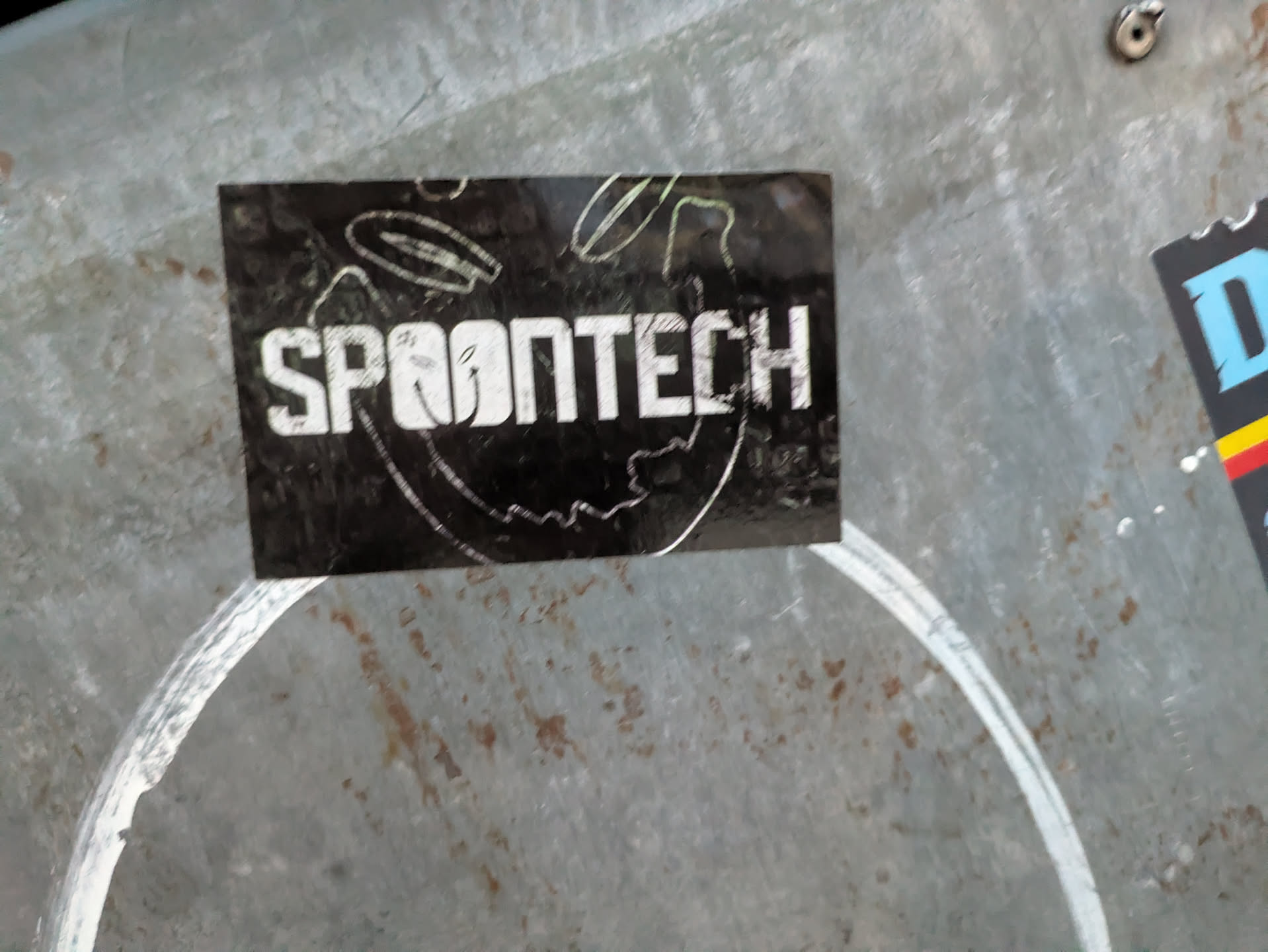 The sticker is rectangular with a black background. The text 'SPOONTECH' is prominently displayed in a bold, white, sans-serif font. The letters have a slightly distressed or worn appearance. There are thin, sketchy white lines and circular shapes around the text, suggesting a dynamic or technical design. The sticker appears to be adhered to a metallic surface with a visible grain and some rust spots.