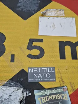 A gray sticker with white text that says "NEJ TILL NATO" (No to NATO) and "FÖR FRED & ALLIANSFRIHET" (For peace and alliance freedom) is affixed to a yellow traffic sign. The sticker is slightly worn and the sign shows other stickers and damage. 