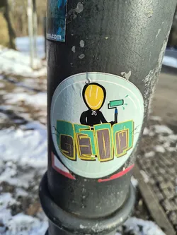 A round sticker is affixed to a dark gray pole. The sticker has a light blue background and a white border. Centered on the sticker is a black figure with a yellow smiley face. The figure holds a paint roller in its hand, with blue paint dripping from the roller. Below the figure, the word "SEDQ" is written in green and yellow block letters.