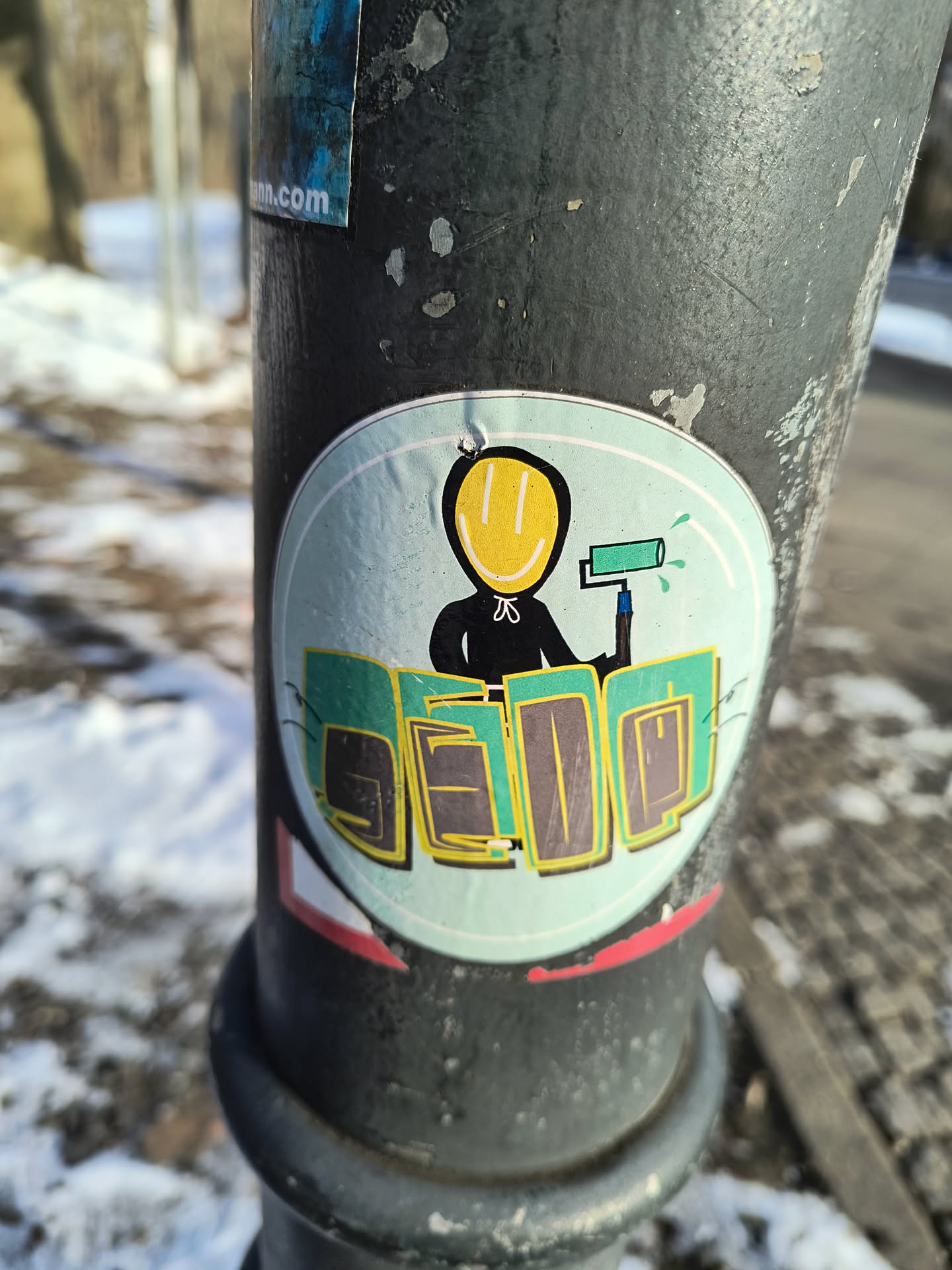 A round sticker is affixed to a dark gray pole. The sticker has a light blue background and a white border. Centered on the sticker is a black figure with a yellow smiley face. The figure holds a paint roller in its hand, with blue paint dripping from the roller. Below the figure, the word "SEDQ" is written in green and yellow block letters.