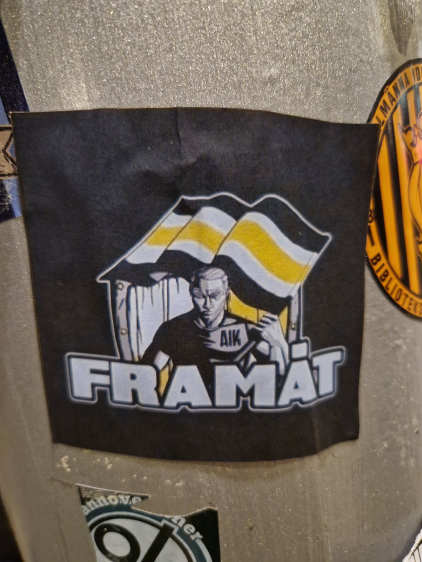 A black and yellow sticker with an illustration of a person holding a flag that says AIK. The text "FRAMÅT" is written underneath the illustration.  The style is reminiscent of political or activist art.