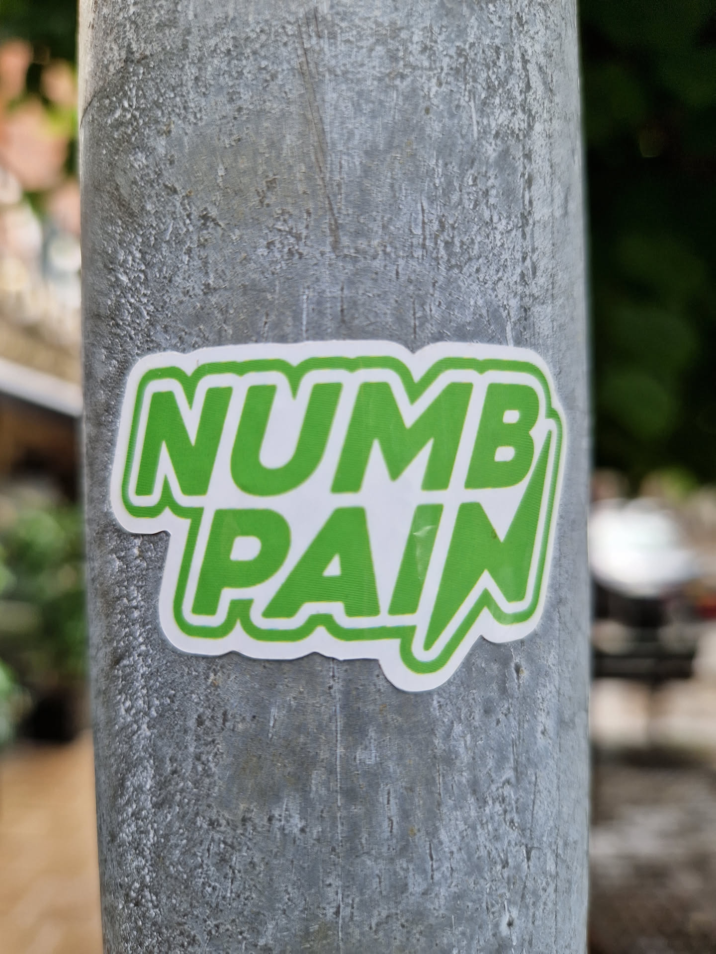 A green and white sticker with the words "NUMB PAIN" in a bold, stylized font. The sticker is affixed to a gray metal pole.