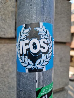 A sticker with the acronym IFOS in a laurel wreath. The sticker is light blue, black, and white. It is affixed to a galvanized metal pole.