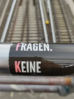A black sticker with white and pink text on a dark surface. The text says "FRAGEN. KEINE" which translates to "QUESTIONS. NO" in German.