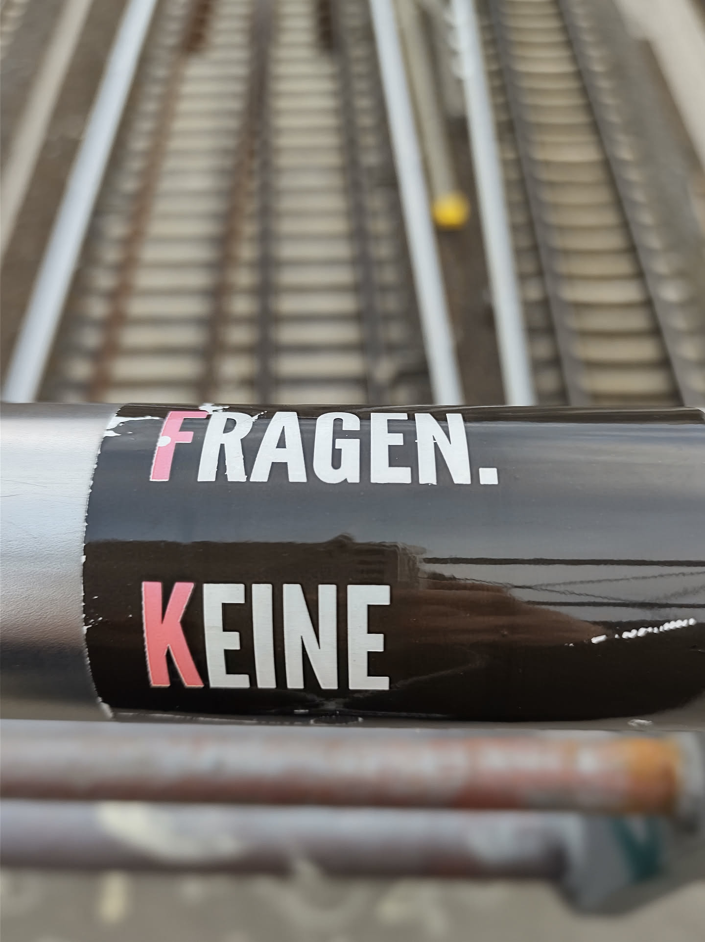 A black sticker with white and pink text on a dark surface. The text says "FRAGEN. KEINE" which translates to "QUESTIONS. NO" in German.