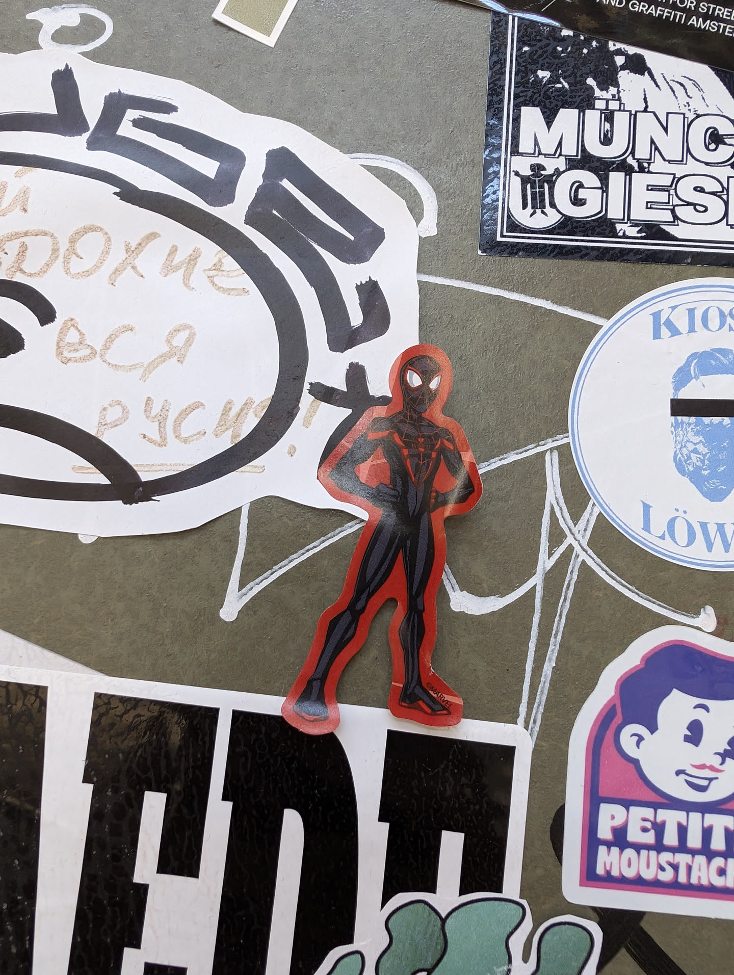 A vinyl sticker featuring a stylized image of Spider-Man in a red and black suit. The sticker is placed on a surface with various other stickers and graffiti.