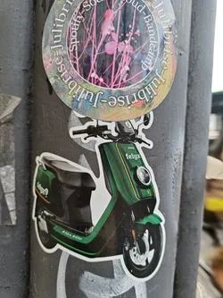 A sticker featuring a green electric scooter, possibly a felqx model.  The sticker is affixed to a pole, surrounded by other stickers and graffiti. One of the visible stickers has the words Spotify, Bandcamp, and 'Julisrise' repeating around a floral design.