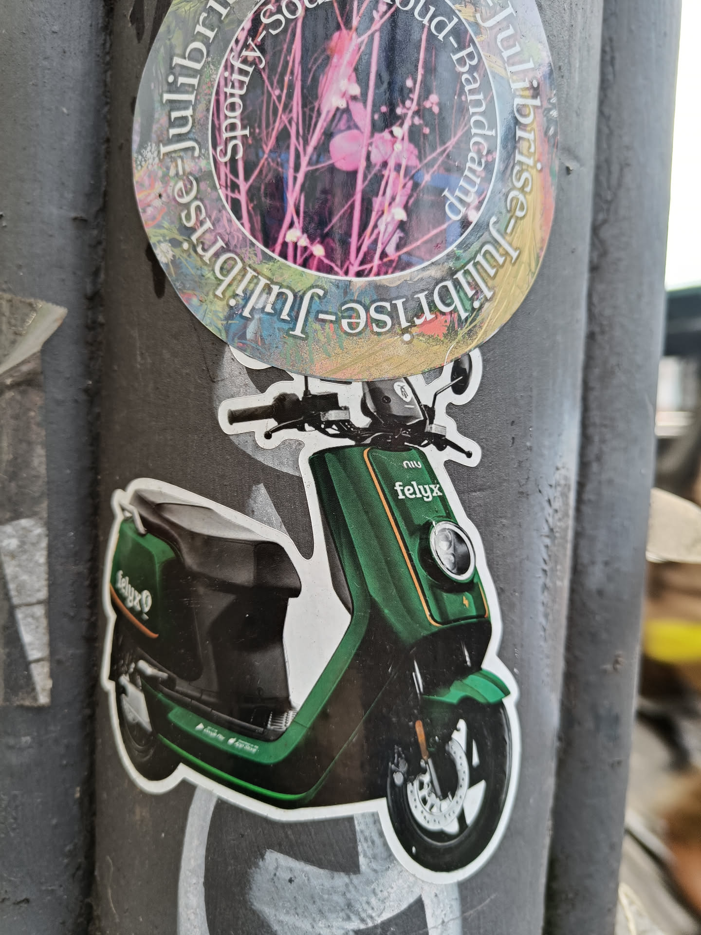 A sticker featuring a green electric scooter, possibly a felqx model.  The sticker is affixed to a pole, surrounded by other stickers and graffiti. One of the visible stickers has the words Spotify, Bandcamp, and 'Julisrise' repeating around a floral design.