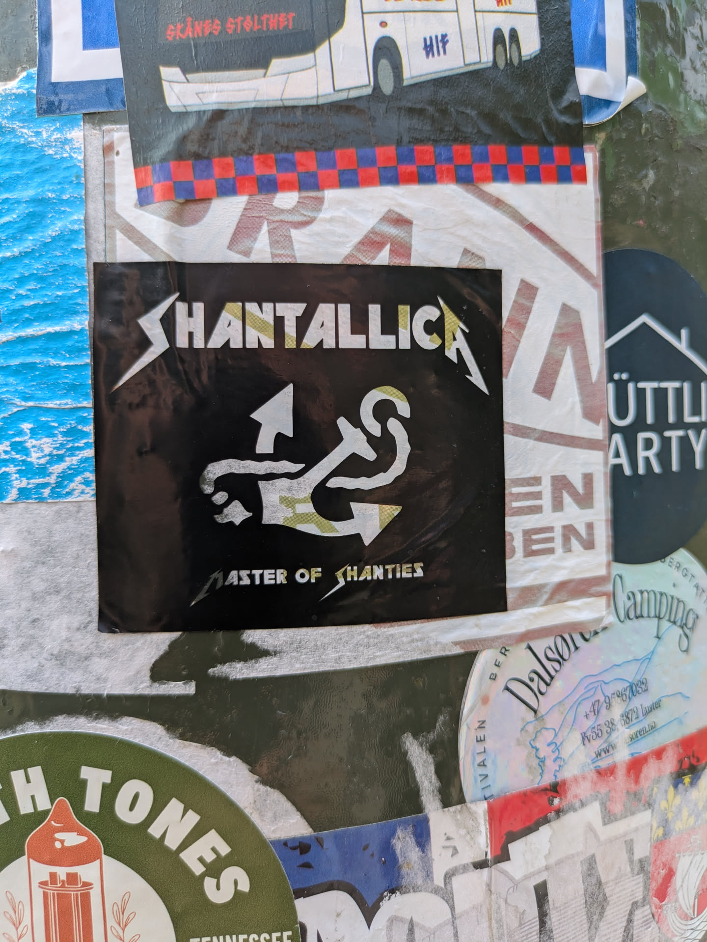 The central sticker is a rectangular black decal. At the top, the word "SHANTALLICA" is prominently displayed in a bold, angular white font, stylistically mimicking the well-known Metallica band logo. The letters 'LLICA' have a subtle light green or camouflage-like texture. Below the band name, there is a distinct graphic symbol, primarily in white with light green accents, which appears to be a creative fusion of an anchor and potentially a musical treble clef or another maritime knot. At the bottom of the sticker, in a smaller, similar font, the phrase "MASTER OF SHANTIES" is written. The design clearly alludes to a blend of heavy metal music and sea shanty themes.