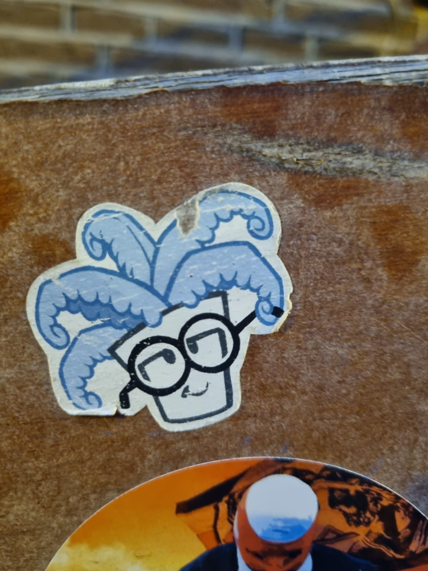 This sticker showcases a charming illustration of a plant with curly tendrils resembling hair, adorned with a pair of spectacles.  The character has a simple, yet expressive face. The sticker itself appears to be made of vinyl and is affixed to a textured, possibly wooden or cardboard surface.