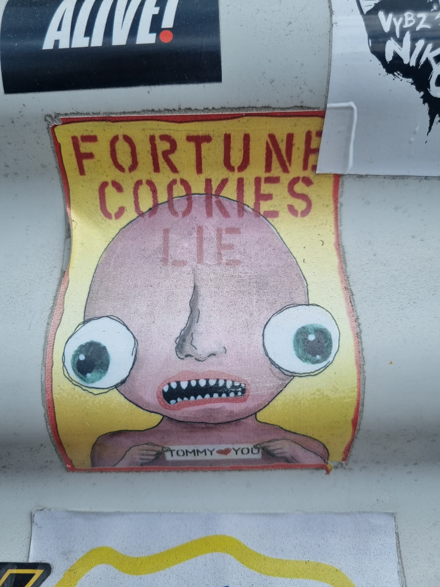 A sticker featuring a cartoon character with large eyes and a simplistic design holding a sign that says "Tommy ♥ Yoo". The background is yellow, and the text "Fortune Cookies Lie" is written at the top.