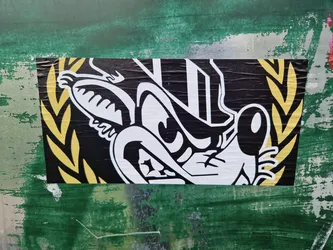 Close-up of a sticker featuring a stylized cartoon character's face. The character is predominantly black and white with a menacing expression. It is framed by a golden yellow laurel wreath. The sticker has a slightly distressed look, suggesting it may be applied outdoors. The background is a textured green surface.