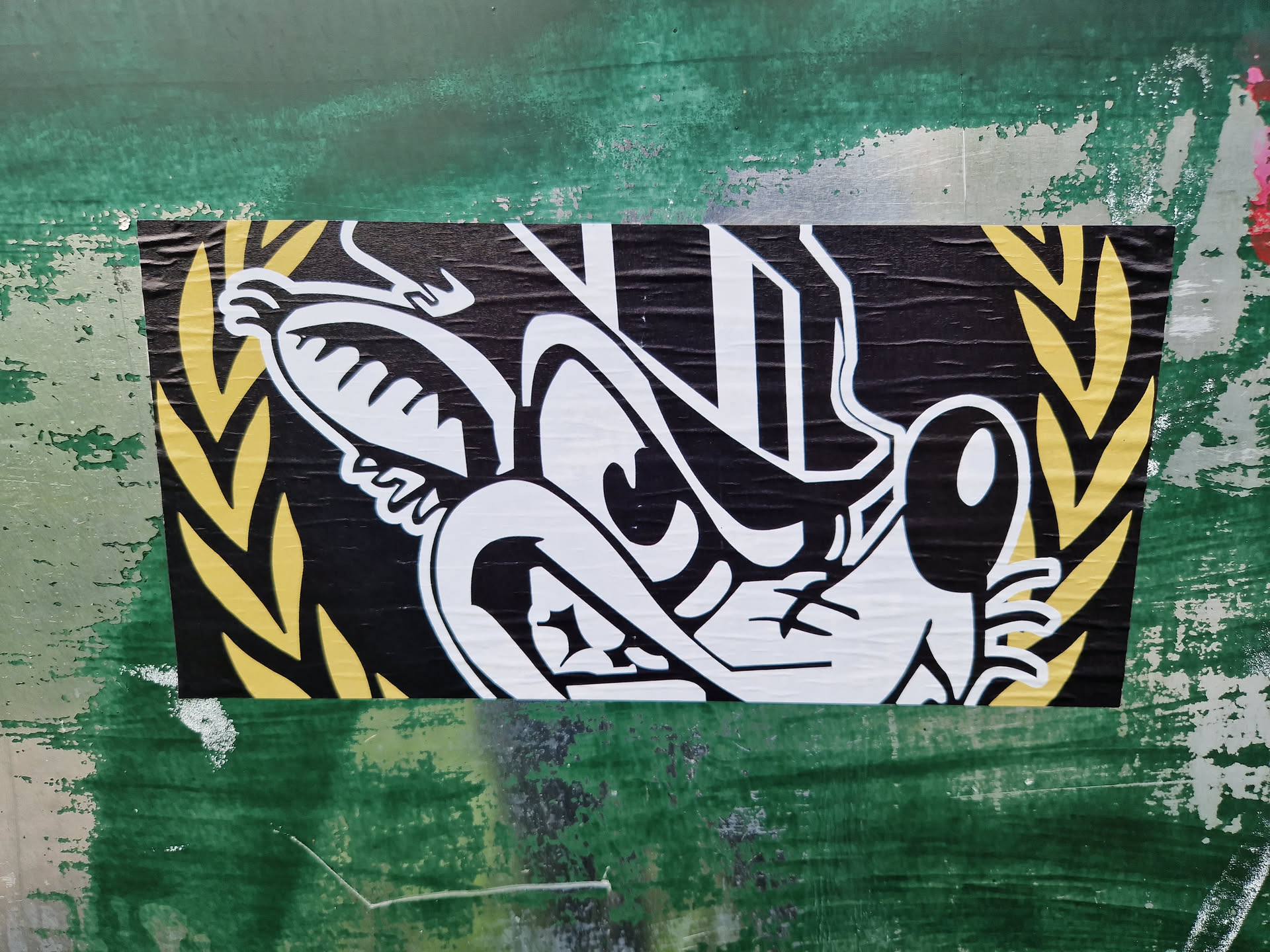 Close-up of a sticker featuring a stylized cartoon character's face. The character is predominantly black and white with a menacing expression. It is framed by a golden yellow laurel wreath. The sticker has a slightly distressed look, suggesting it may be applied outdoors. The background is a textured green surface.