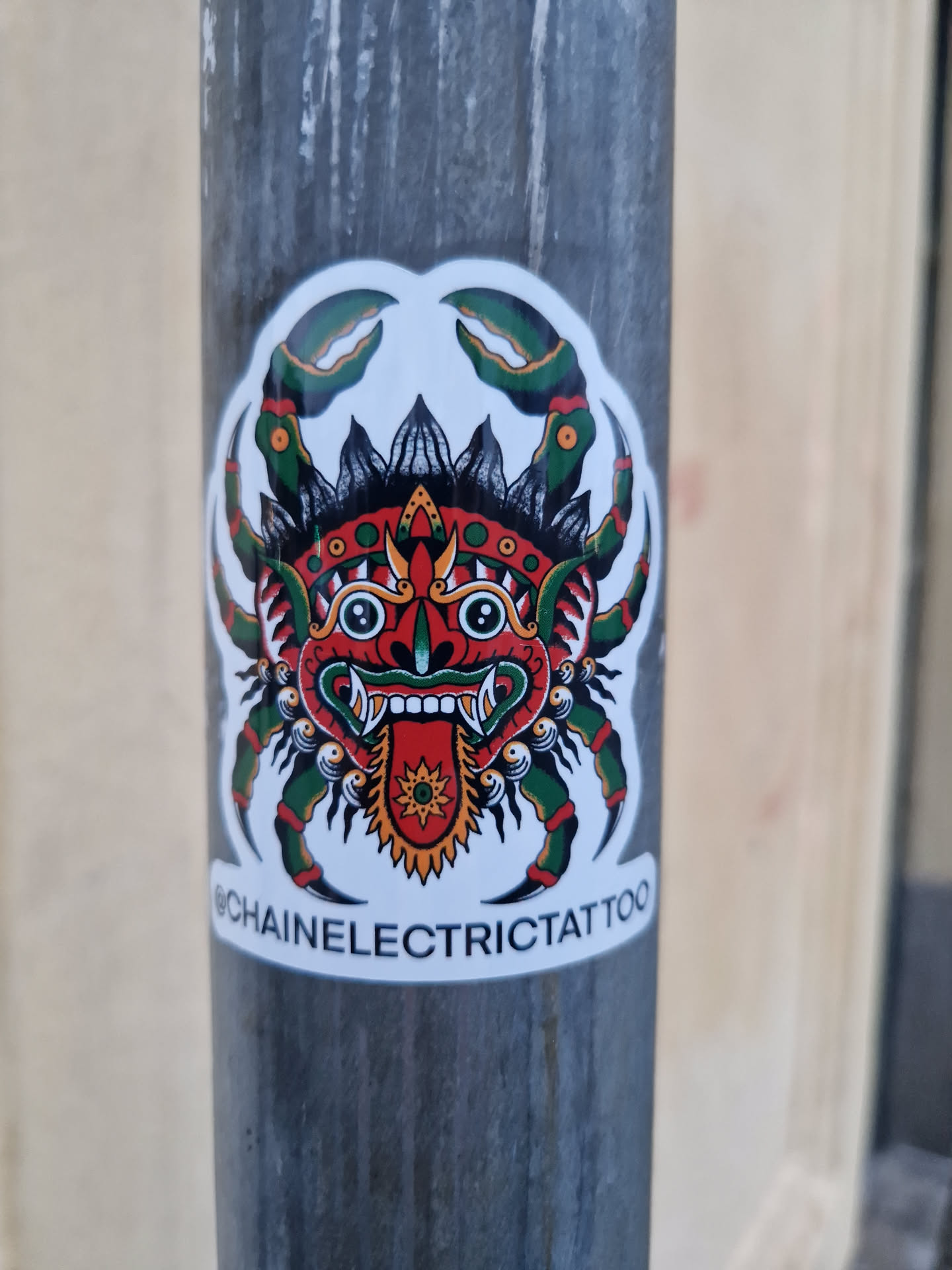 A sticker featuring a traditional tattoo-style illustration of a crab with a demonic face in the center. The crab is depicted with green claws and a red, detailed face. Below the illustration, the text "@chainelectrictattoo" is visible.