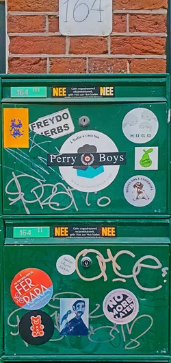 A green mailbox is adorned with multiple stickers of different styles, shapes, and sizes. The stickers range from food advertisements to political statements. There are also handwritten tags on the mailbox.