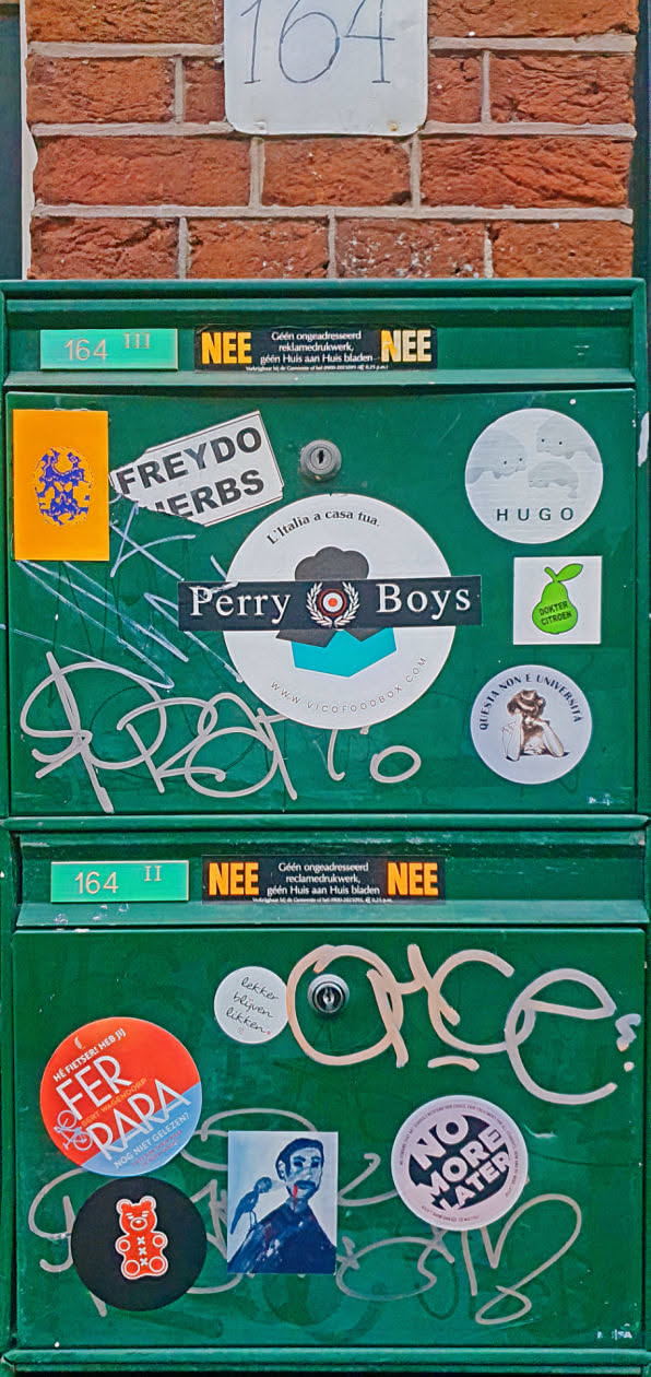 A green mailbox is adorned with multiple stickers of different styles, shapes, and sizes. The stickers range from food advertisements to political statements. There are also handwritten tags on the mailbox.