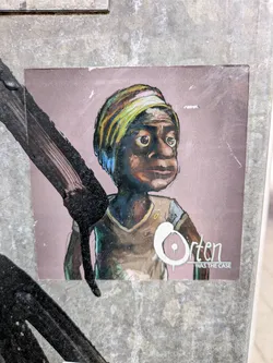 The sticker depicts a cartoonish person with dark skin, a yellow and green head wrap, wearing a beige shirt. The background is a muted mauve color. The text on the sticker says "Orten WAS THE CASE".