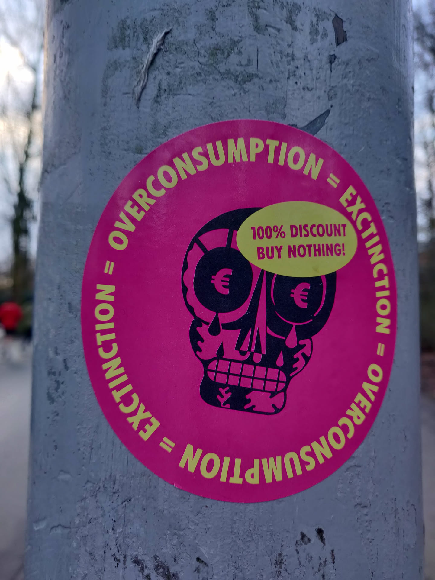 Pink circular sticker with a skull graphic and text. The text on the sticker says: 'Overconsumption = Extinction' and '100% Discount Buy Nothing!' 